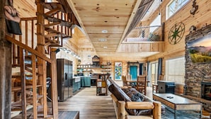 A spacious wooden interior with a central seating area, a kitchen in the background, and a stone fireplace. - Neverland (Gatlinburg)