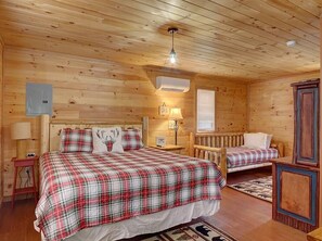 A cabin-style room with a large bed, a smaller bed, a wooden ceiling, a nightstand, a lamp, a dresser, and a window with blinds. - A Family Tradition (Gatlinburg)