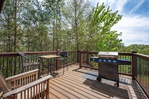A wooden deck with a grill, a table, and chairs, surrounded by trees. - Fox n' Socks (Sevierville)
