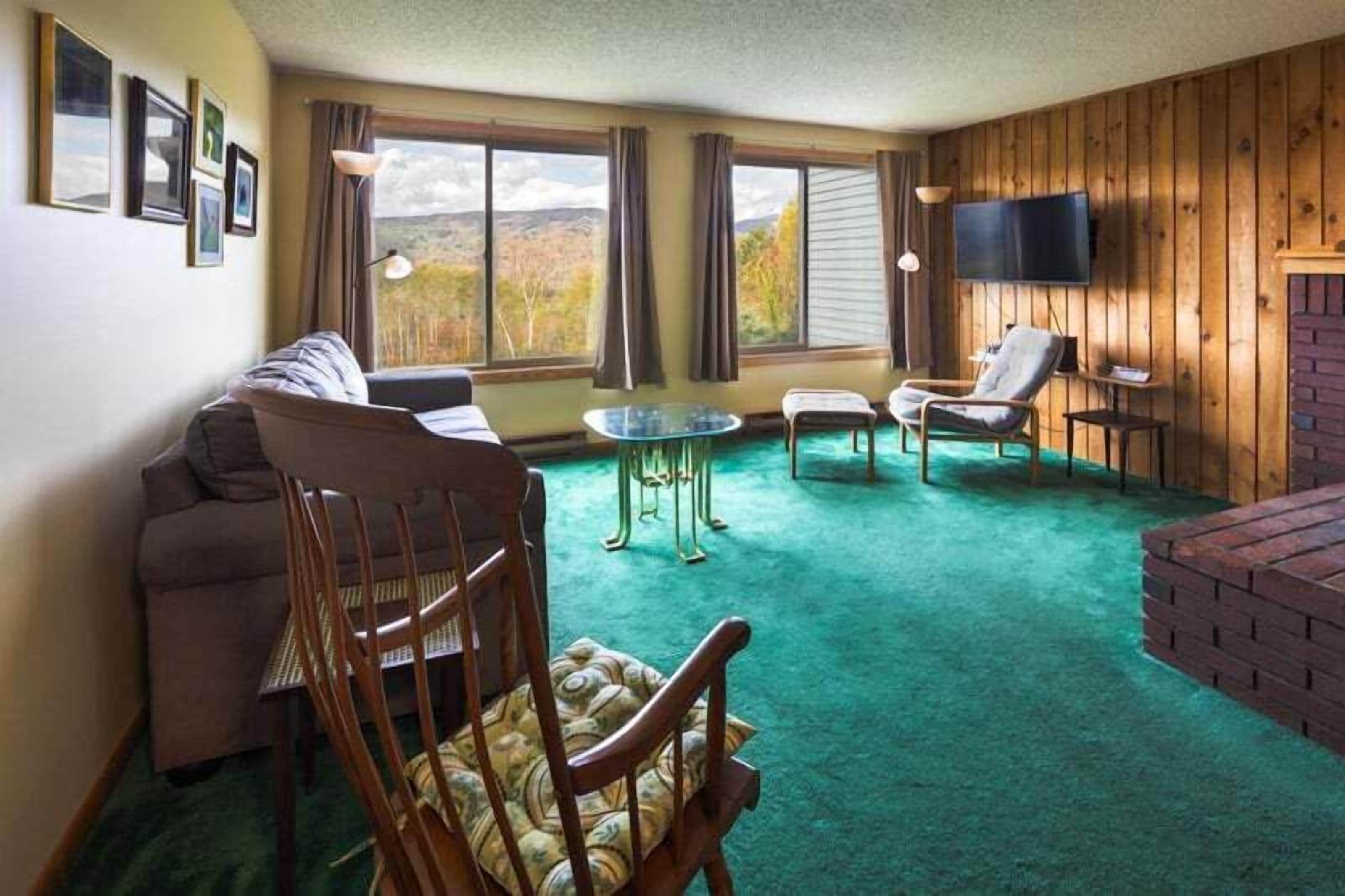 A room with a green carpet, wooden walls, a sofa, a chair, a small table, and a TV mounted on the wall.