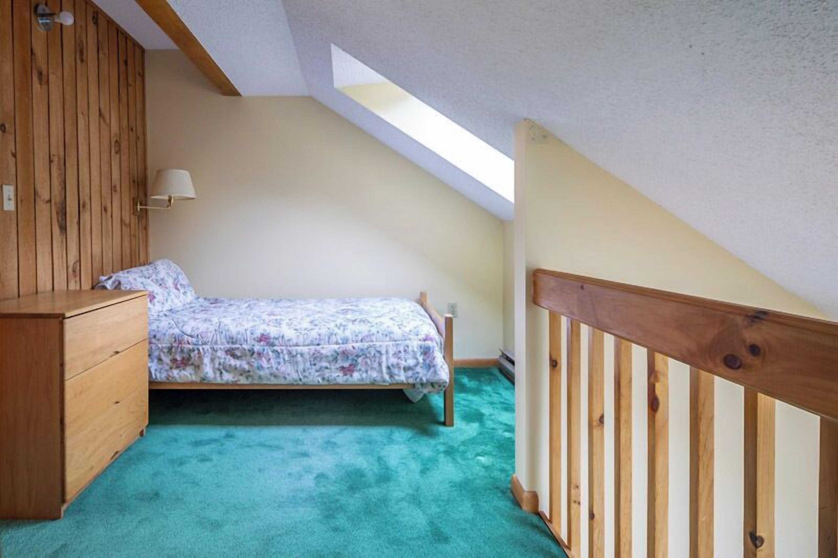 Un dormitorio con techo inclinado, cama, cómoda de madera y tragaluz.