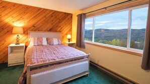 A bedroom with a bed, a nightstand, a window with a view, and a wooden ceiling. - Enjoy stunning view in Waterville Estates! (Thornton)