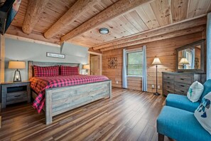 A bedroom with a wooden ceiling, a bed with a checkered bedspread, a nightstand with a lamp, a dresser, and a blue sofa. - Rustic Retreat (Gatlinburg)