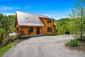 A log cabin with a metal roof, a porch with seating, and a driveway leading to the entrance. - Rustic Retreat (Gatlinburg)