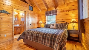 Interior de uma cabana de madeira com uma cama, luminárias de cabeceira, uma janela com cortinas e um pequeno armário.