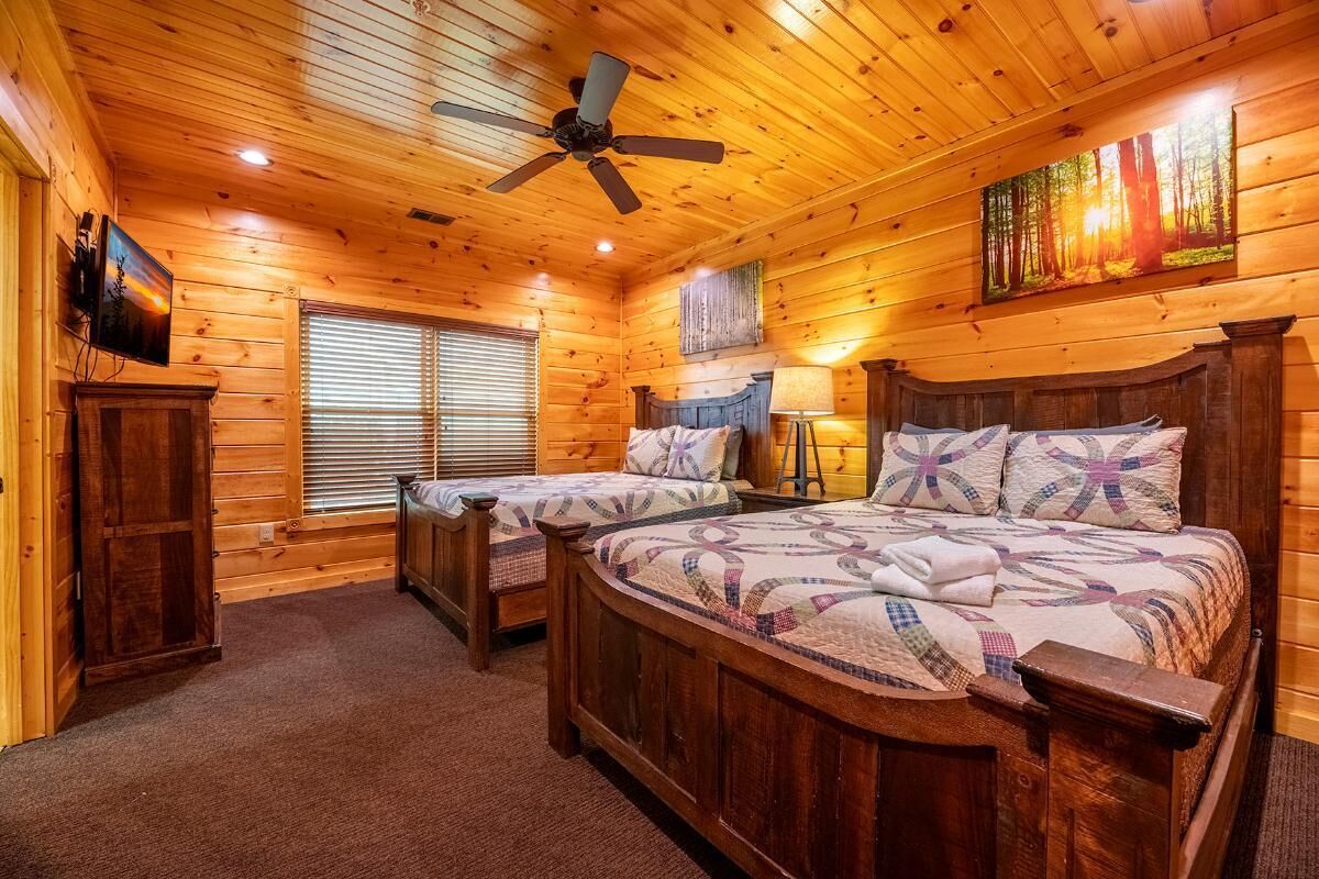 Quarto de cabana de tronco com duas camas, um ventilador de teto, uma TV e um quadro.