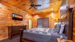 A wooden cabin bedroom with a bed, a dresser, a television, and a ceiling fan. - Mayes Landing (Pigeon Forge)
