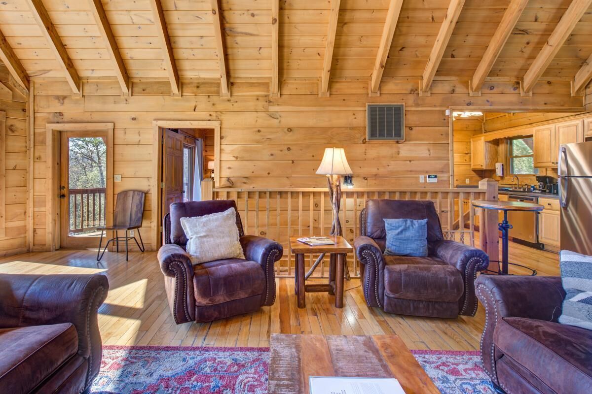 Interior de una cabaña de troncos con una sala de estar que incluye sofás de cuero, una mesa de comedor de madera y una cocina visible a través de un arco.