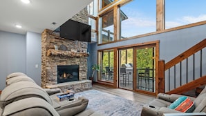 A living room with a stone fireplace, a large window, and a staircase. - Riverside Retreat in Ellijay GA (Ellijay)