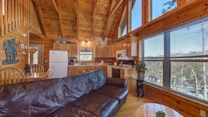 Interior de una cabaña de madera con sofá de cuero, cocina, comedor y ventanas amplias.