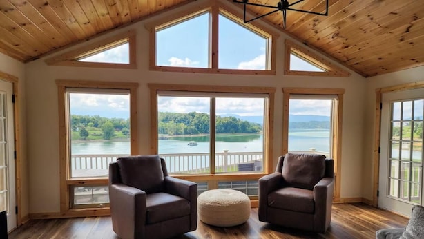 A room with wooden flooring, two leather armchairs, a round ottoman, and large windows with a view of a lake and trees. - Three Horizons Retreat (Dandridge)
