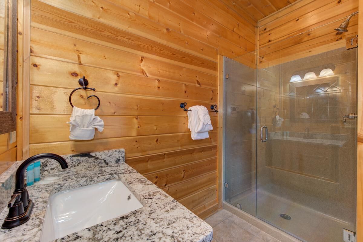 Une salle de bain rustique, avec des murs en bois, un lavabo sur un plan de travail en granit, une douche en verre et des serviettes suspendues à des crochets.