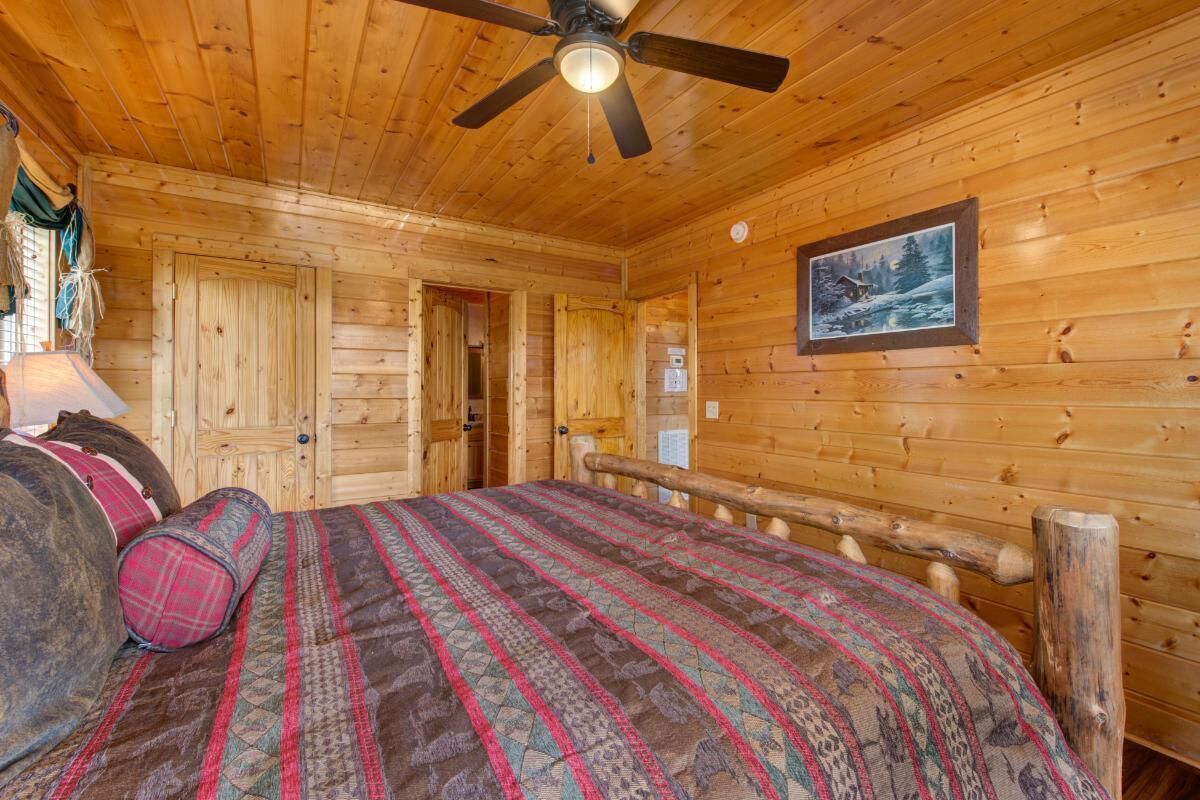 A wooden cabin room with a bed, a ceiling fan, a door, and a framed picture on the wall.
