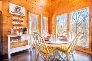 A wooden cabin interior with a dining table, chairs, a coffee station, and a view of trees through large windows. - Smoky Summit Stays (Sevierville)