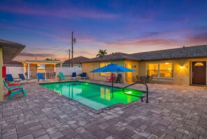 Une piscine entourée d’une terrasse en briques, avec des chaises longues et un parasol.