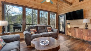 A wooden cabin interior with a sofa, a coffee table, a TV, and large windows. - Bearfoot Splash Hideaway (Sevierville)