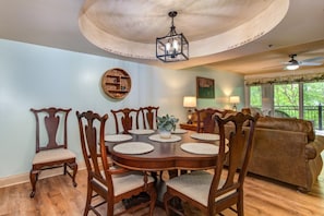 A dining area with a round wooden table, six chairs, a chandelier, a wall-mounted shelf, and a window with a view of greenery. - Cs Creekside Condo (Gatlinburg)