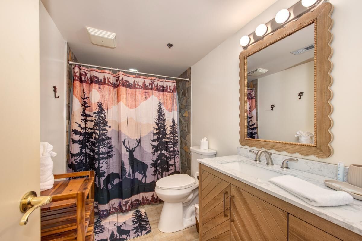 Un baño con cortina de ducha con diseño de bosque y ciervos, un inodoro blanco, un tocador con espejo y un perchero para toallas.