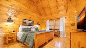 A wooden cabin bedroom with a bed, bedside table, lamp, painting, and a TV. - Roudys Roost (Pigeon Forge)