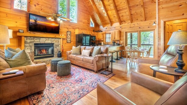 A log cabin interior with a stone fireplace, leather sofas, a television, and a dining area. - Roudys Roost (Pigeon Forge)