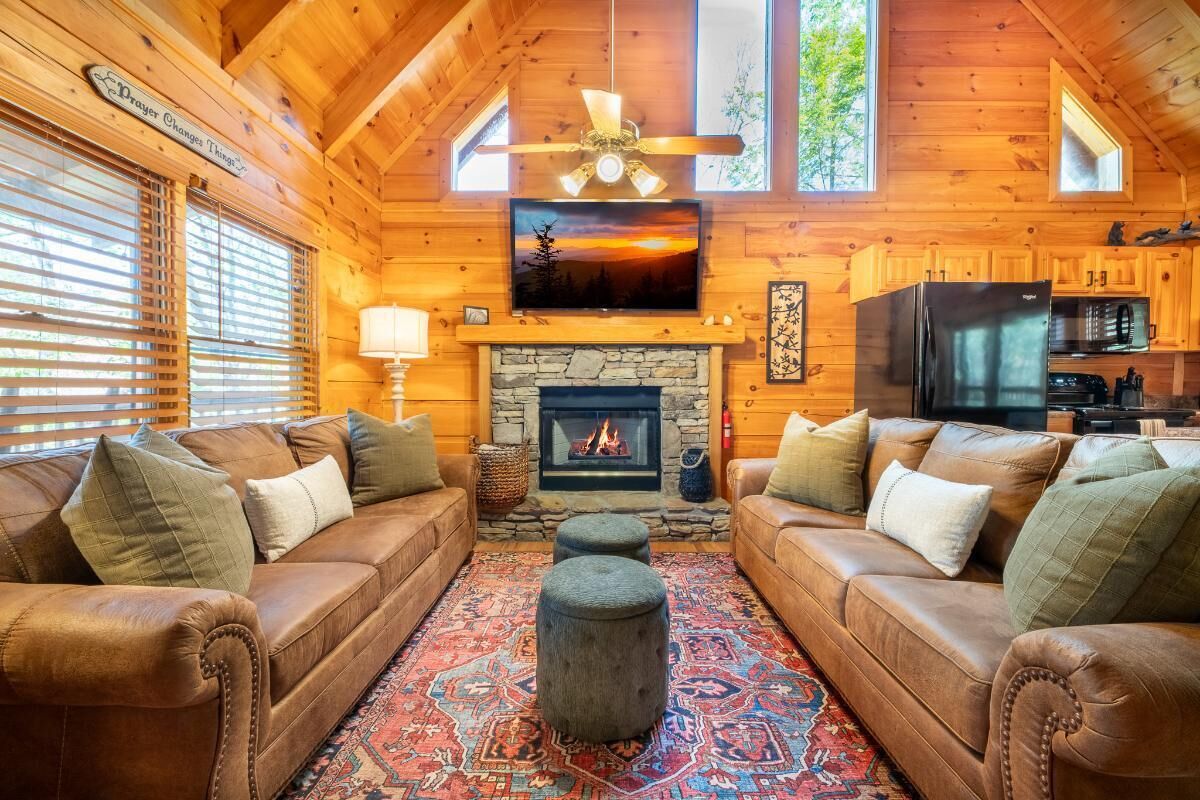 A cozy log cabin interior with a fireplace, leather sofas, a television, and a stone fireplace surround.