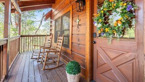 A wooden porch with rocking chairs, a potted plant, and a door with a floral wreath. - Roudys Roost (Pigeon Forge)
