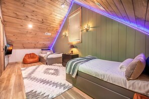 Ein gemütliches Schlafzimmer im Dachgeschoss mit schragem Dach, Holzbalken, einem Bett mit Kissen, einem Nachttisch, einer Lampe und einem Sitzsack.