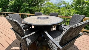 Une table ronde d’extérieur, entourée de six chaises en osier, est posée sur une terrasse en bois, elle-même entourée d’arbres.