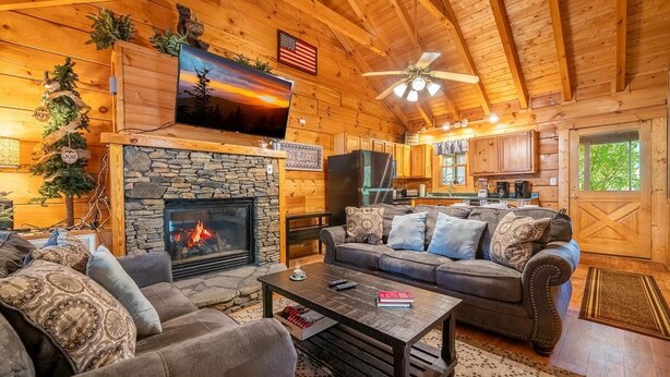 L’intérieur chaleureux d’un chalet comprend une cheminée en pierre, un téléviseur fixé au-dessus, un coin repas et une cuisine avec des armoires en bois.