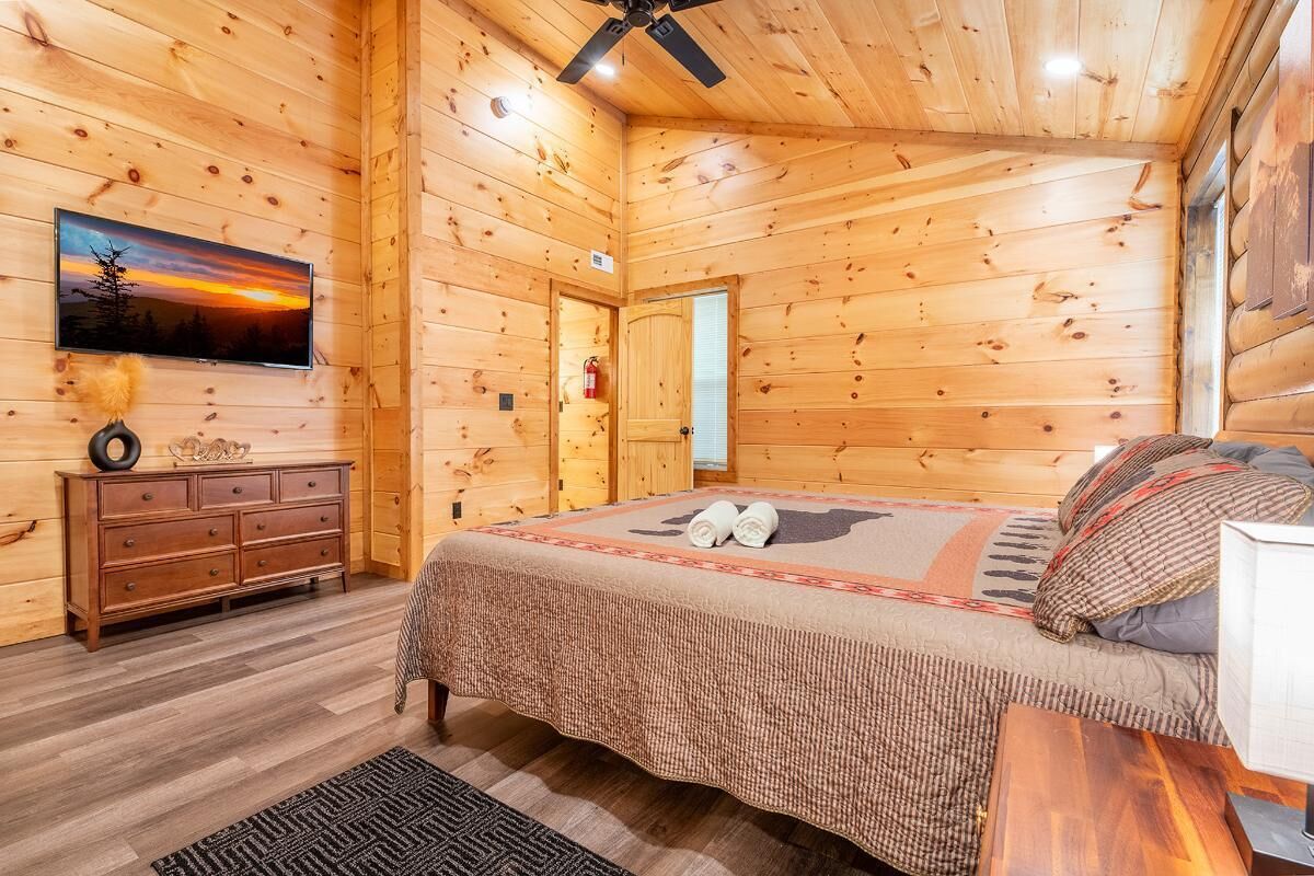 A wooden cabin bedroom with a bed, dresser, television, and a ceiling fan.