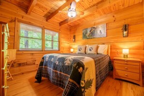 Quarto de cabana de madeira com uma cama, mesas de cabeceira, um ventilador de teto e um quadro na parede.