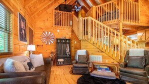 Ein Interieur einer Holzhütte mit Treppe, Kamin, Ledersofas und einem schwarzen, flauschigen Teppich.