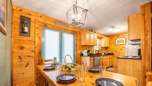 L’intérieur d’un chalet en bois comprend une table à manger pour deux personnes, un coin cuisine avec des placards en bois et un luminaire suspendu.