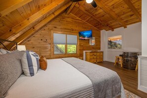 A bedroom with a wooden ceiling, a bed with pillows, a TV, a window with a view, a wooden dresser, and a barrel. - Timber Trace (Pigeon Forge)