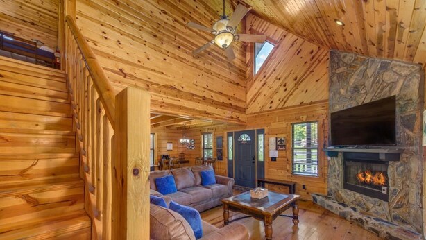 A log cabin interior with a stone fireplace, wooden floors, a ceiling fan, and a large TV mounted on the wall. - Goldilocks and The Three Bears (Gatlinburg)