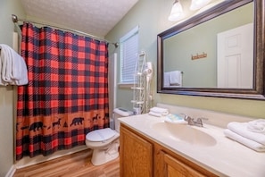 A bathroom with a checkered shower curtain, a toilet, a sink with a mirror, and a window with blinds. - Z - Four Cubs Cottage (Sevierville)