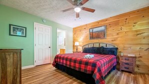 Ein Schlafzimmer mit einem hölzernen Kopfteil, einem Bett mit rot-schwarz karierter Bettdecke, einem Deckenventilator, einem Nachttisch aus Holz mit einer Lampe und einer Tür.