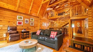 L’intérieur d’une cabane en rondins, avec un escalier en bois, un canapé, un fauteuil et des tableaux encadrés accrochés aux murs.