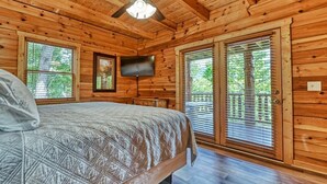 A wooden cabin bedroom with a bed, a ceiling fan, a TV, and large windows. - Mountain Song (Gatlinburg)