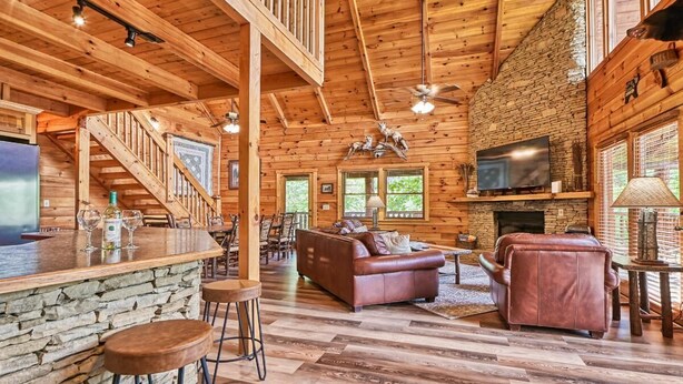 A log cabin interior with a stone fireplace, wooden floors, a bar area, and a cozy seating arrangement. - Mountain Song (Gatlinburg)