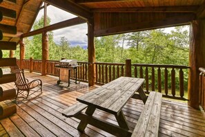 A wooden deck with picnic tables and a grill, surrounded by a wooden railing with a view of trees. - Mountain Song (Gatlinburg)