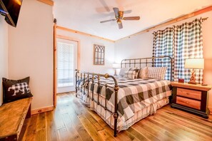 A bedroom with a bed, wooden floor, a ceiling fan, a window with blinds, a bedside table with a lamp, and a checkered bedspread. - Oldham Ridge Retreat (Sevierville)