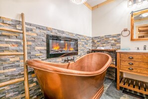A bathroom with a stone wall, a fireplace, a copper bathtub, and a wooden vanity. - Oldham Ridge Retreat (Sevierville)
