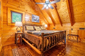 A log cabin bedroom with a wooden bed, a ceiling fan, a window with blinds, and a framed picture on the wall. - Sharp Dressed Bear (Sevierville)