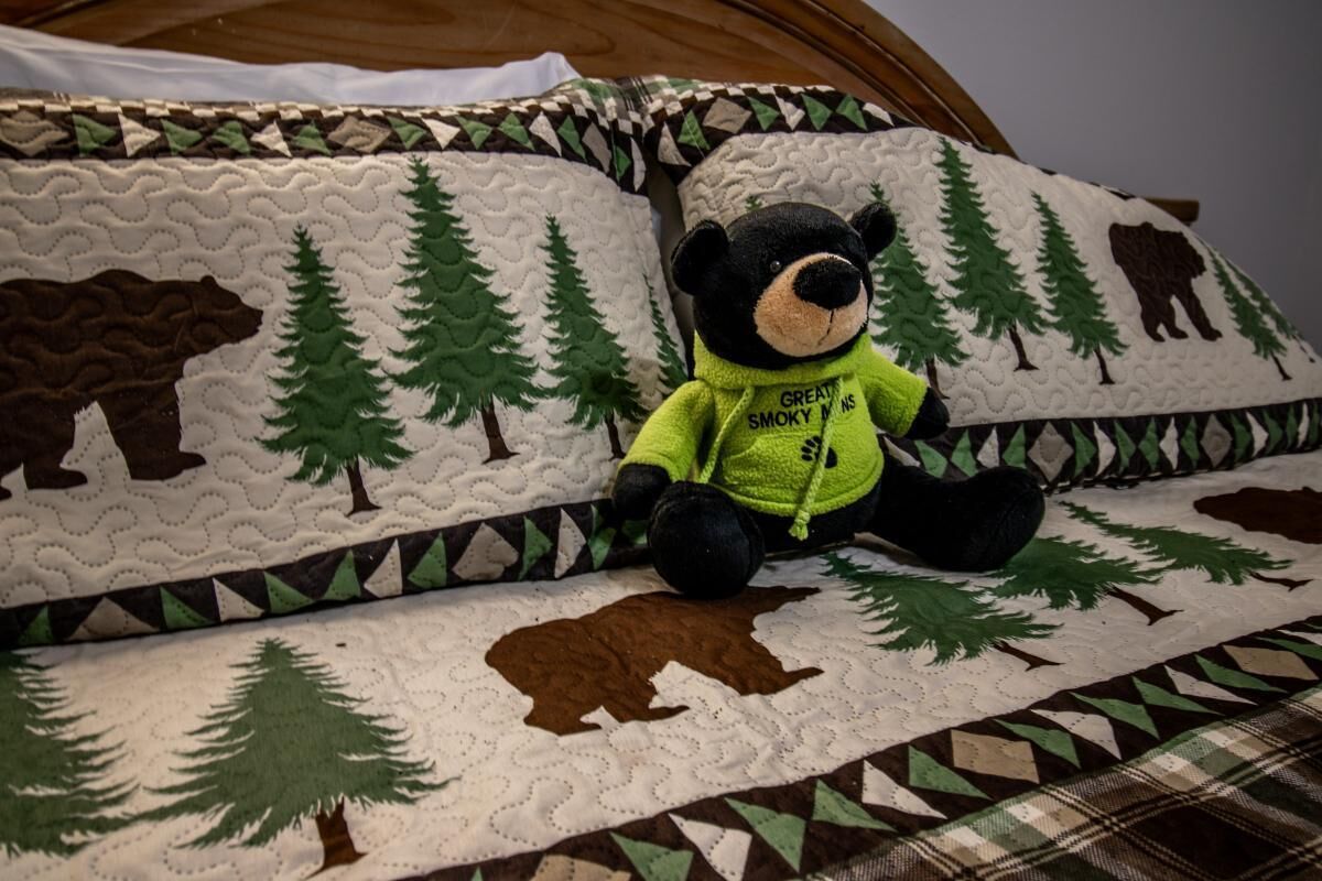 Ein Bett mit einer gesteppten Decke mit Bären- und Baum-Motiv, einem Plüschbären und einem Kopfkissen im gleichen Design.