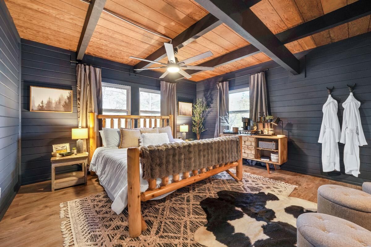 Ein Schlafzimmer mit einem h ölzernen Bett, einem Deckenventilator, einem Nachttisch, einer Lampe, einem Fenster und einem Kuhfellteppich.