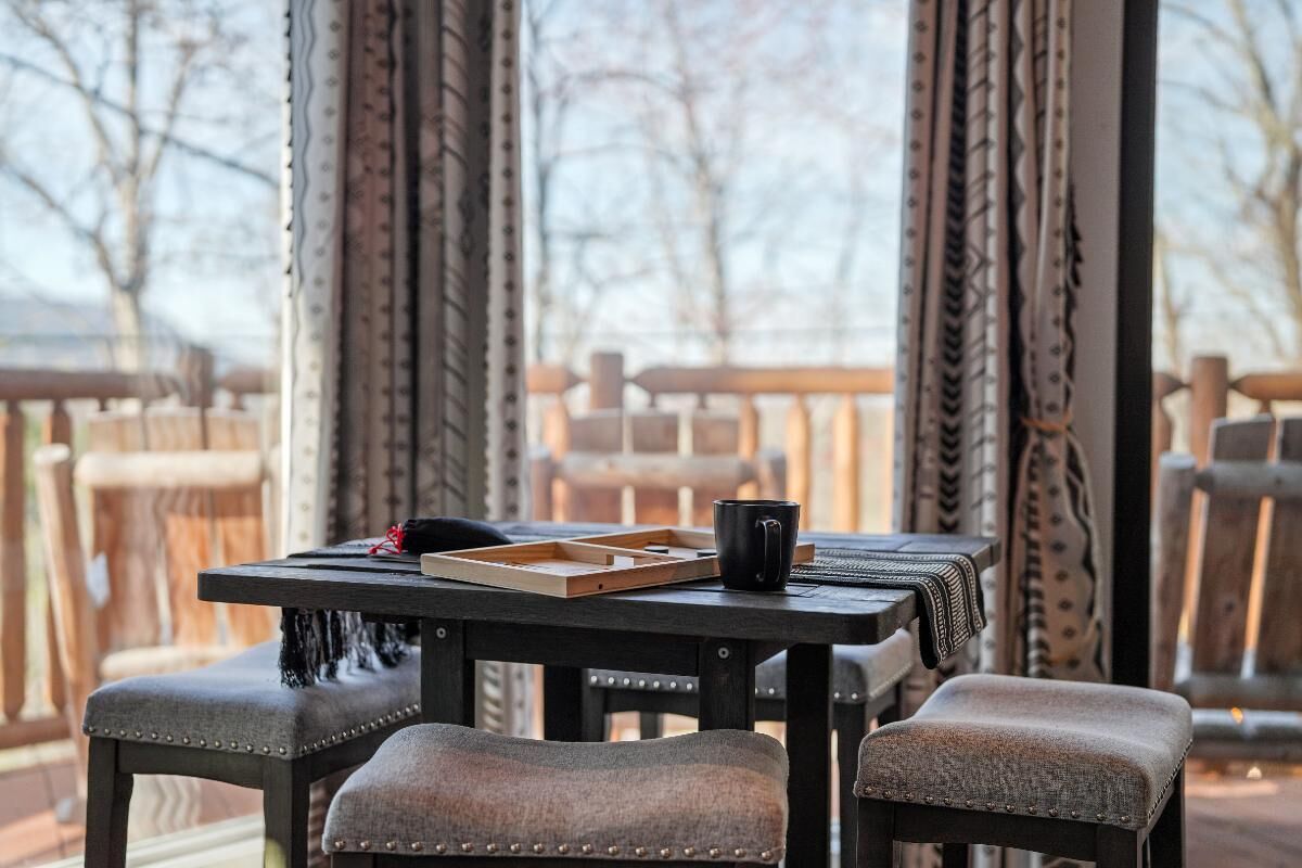 Une table avec un plateau, une tasse noire et un tapis tissé sur un balcon agrémenté de rideaux à motifs et d’une rambarde en bois.