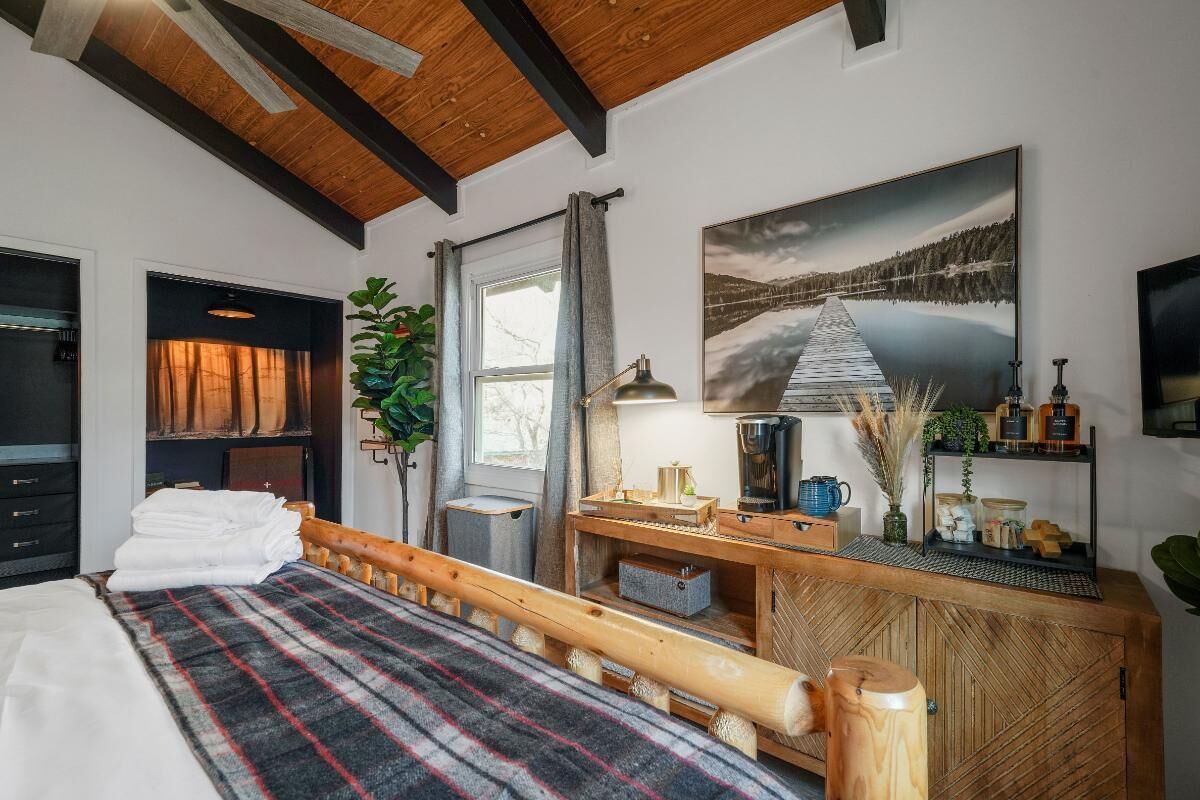 Ein Schlafzimmer mit einem Bett, einem Holzkopfstück, einem Schreibtisch mit Lampe, einem Gemälde und einem Fenster mit Vorhängen.