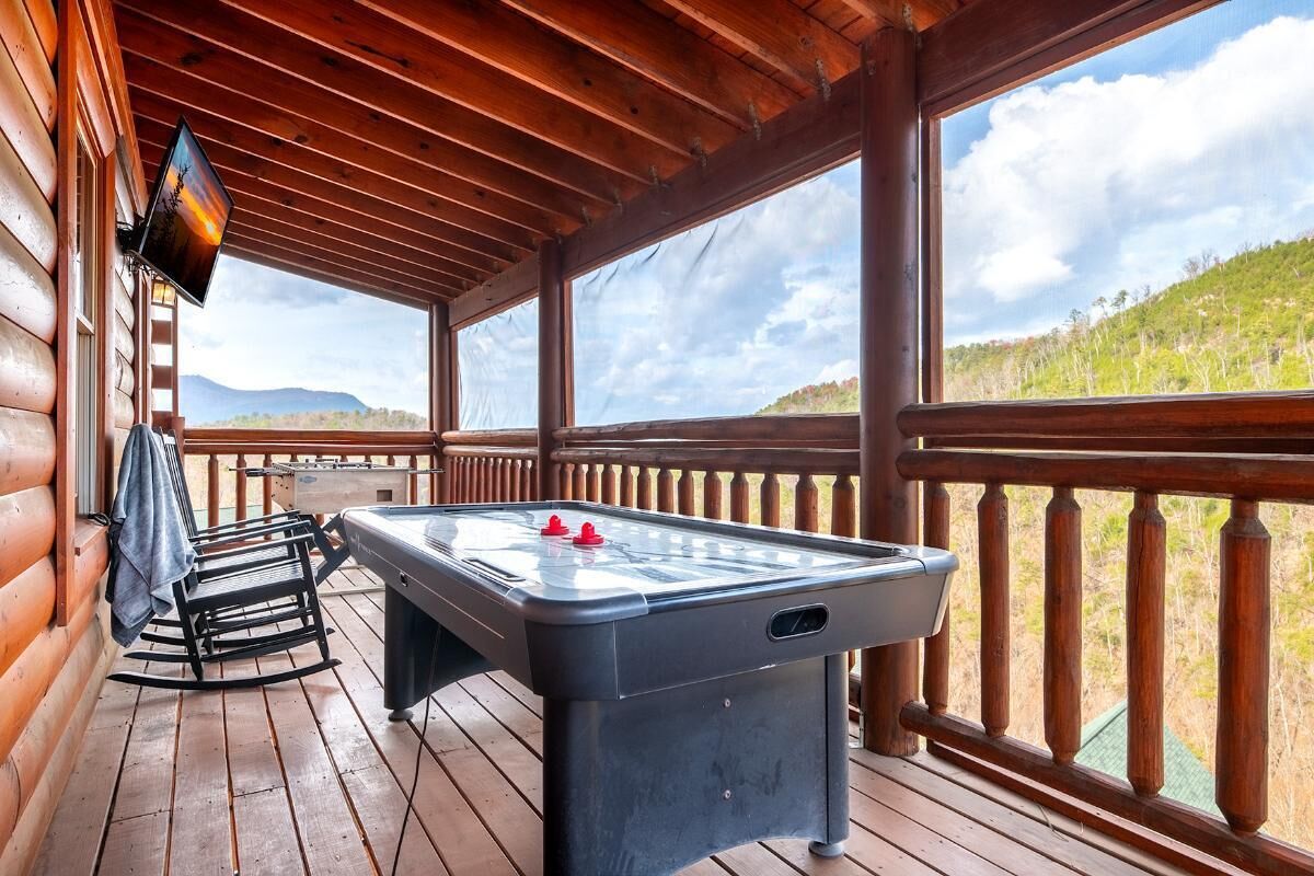 A wooden deck with a pool table, a TV mounted on the wall, and a view of a hilly landscape.