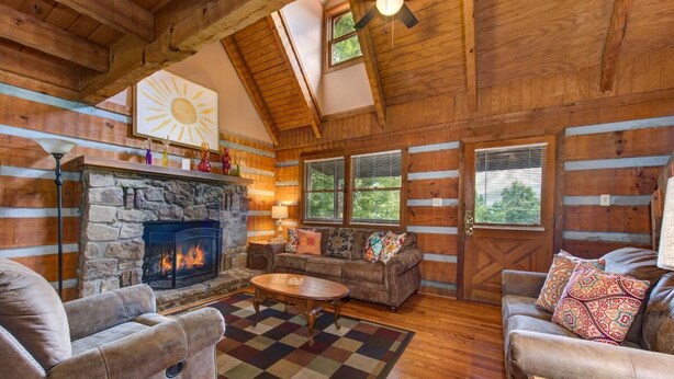 A log cabin interior with a stone fireplace, wooden ceiling beams, a ceiling fan, and a large window. - Paradise (Gatlinburg)